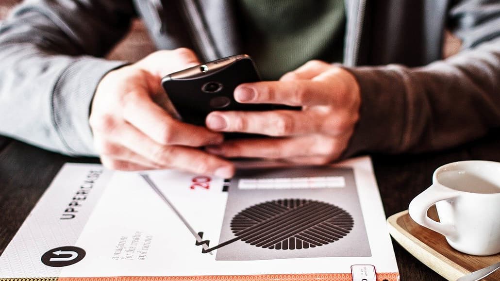 Man using an android phone with two hands resting on a table with a book and coffee cup